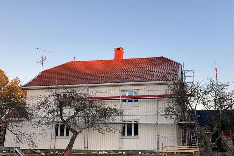 Et hus med rød takstein og stillas, under oppussing, med trær i forgrunnen og klar himmel i bakgrunnen.