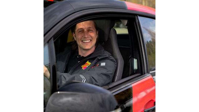 En smilende mann sitter i førersetet i en bil med svart og rød lakk.