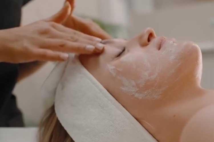 En kvinne får ansiktsbehandling med ansiktsmaske på et spa, mens en terapeut masserer pannen hennes.