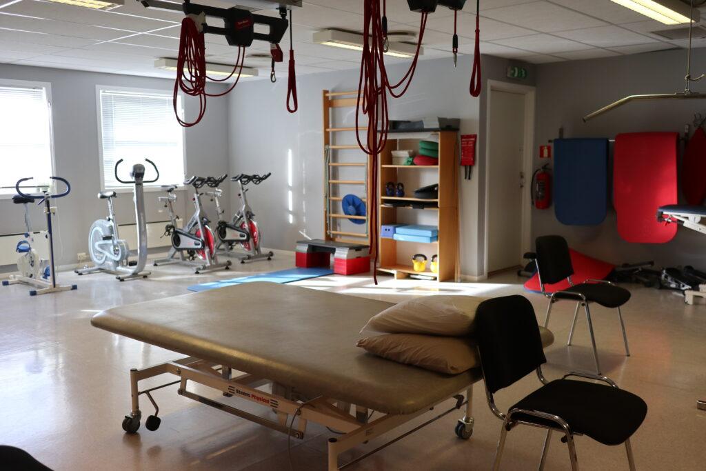 Treningsrom med treningsutstyr, en behandlingsbenk med puter og stoler, samt lys fra vinduer.