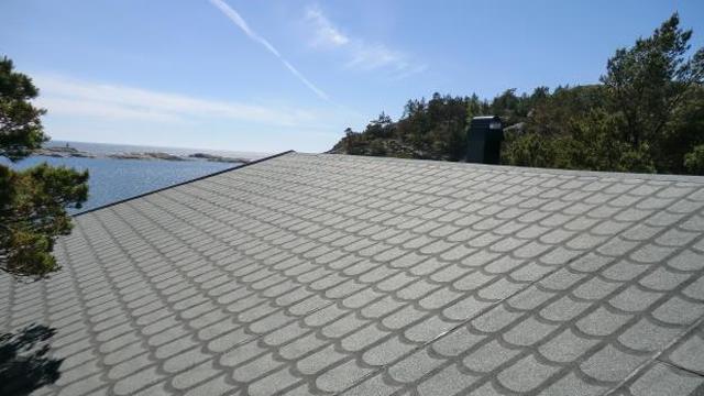 Tak med grå takstein og utsikt over havet og trær i bakgrunnen på en solfylt dag.