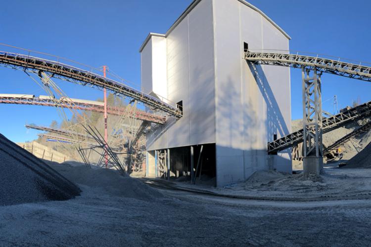 En stor gruvebygning med transportbånd som frakter stein og grus i et steinbrudd under klar himmel.