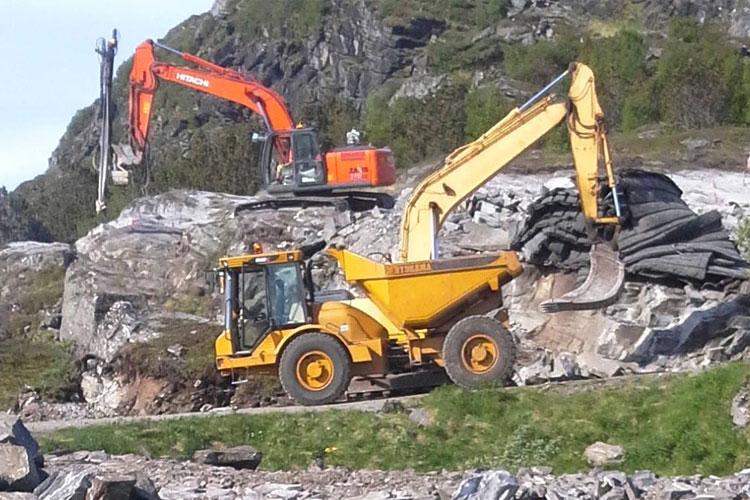 To store og gule gravemaskiner arbeider på en steinete byggeplass med fjell i bakgrunnen.