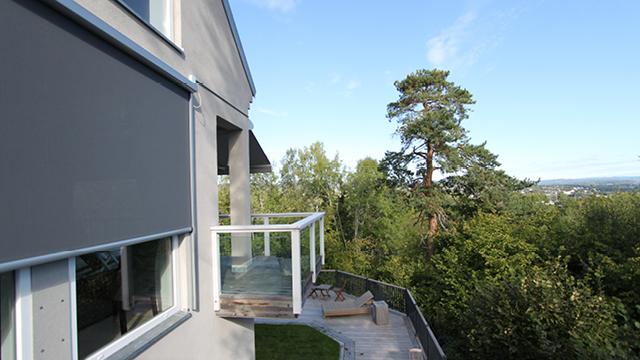 Moderne hus med terrasse og utsikt over skog og landskap, omkranset av trær og blå himmel.
