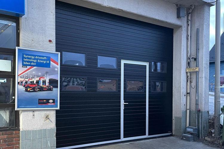 Svart garasjeport med glassvinduer og en dør, plassert på en bygning med reklameskilt ved siden av.