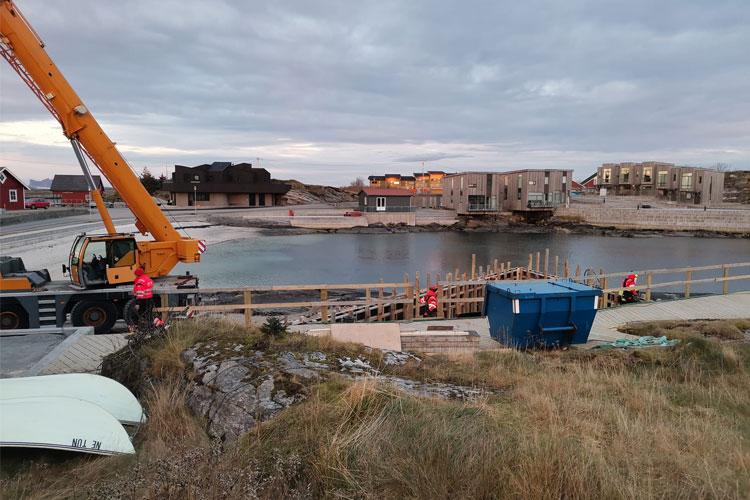 Kran som arbeider ved en båtkaip, med bygninger og en blå container i bakgrunnen.