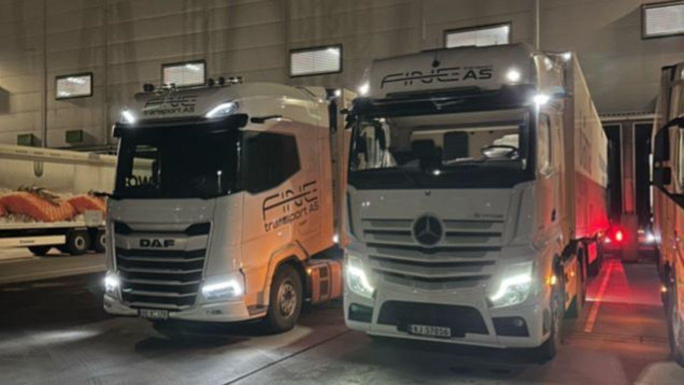 To lastebiler parkert i en lagerbygning, en DAF og en Mercedes, begge med lys på.