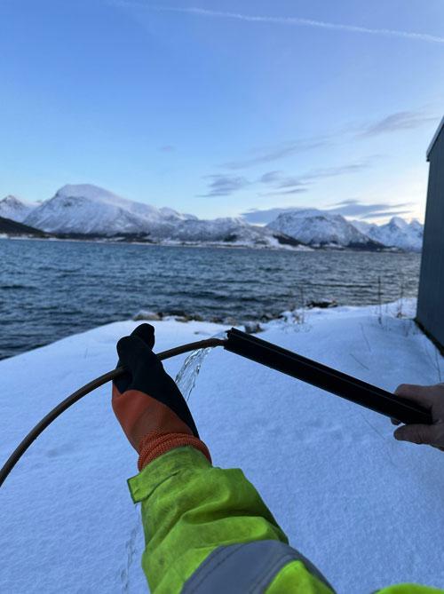 En person i refleksjakke holder en slange over snøen ved vannkanten, med fjell i bakgrunnen.