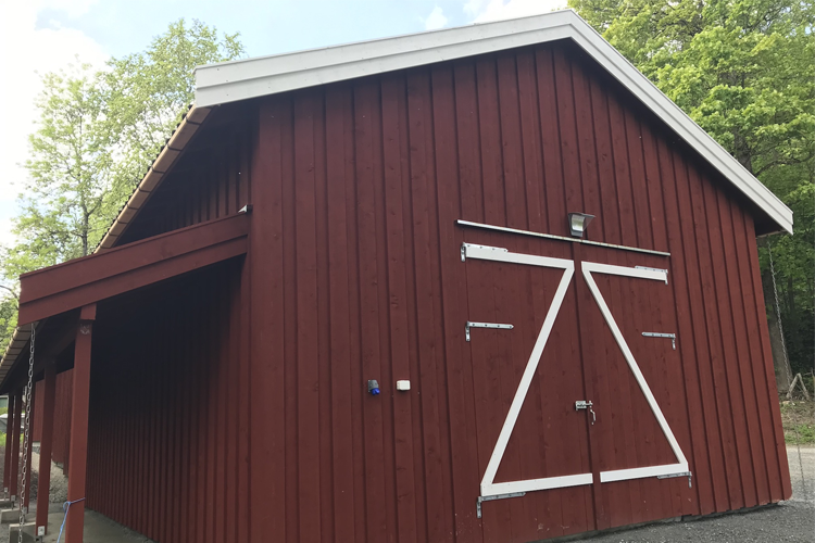 Rød låve med hvite detaljer og skråtak, omringet av grønn vegetasjon.