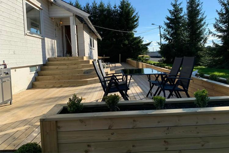 Terrasse med treplanker, stoler og planter, solfylt med trær i bakgrunnen.