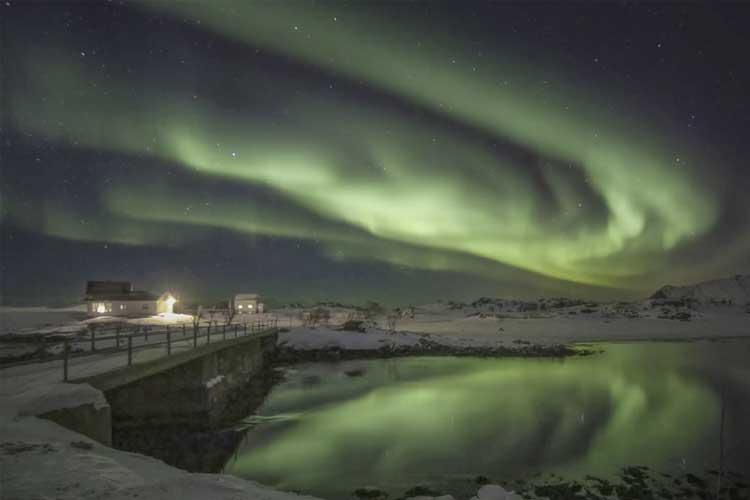 Nordlys over en stille innsjø med snødekte fjell og en bro i forgrunnen. Huset lyser opp i natten.