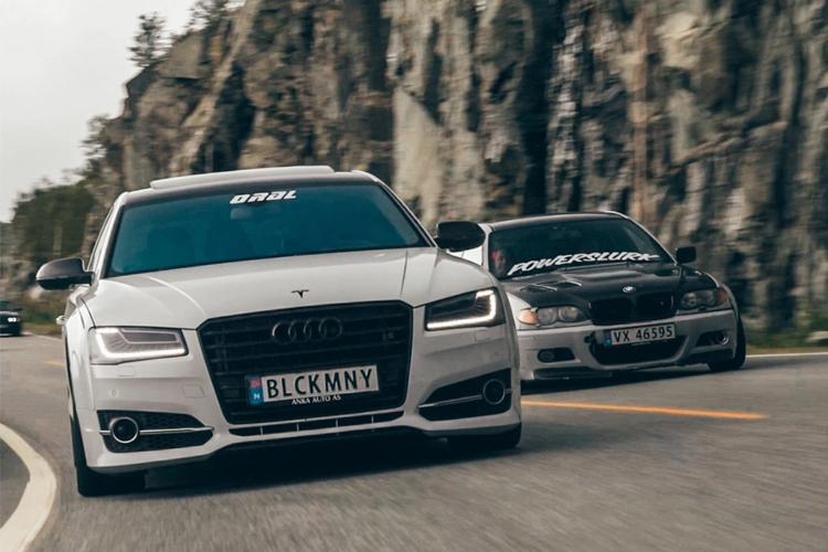To biler som kjører på en svingete vei, en sølvfarget Audi A8 og en mørk bil med klistremerker.