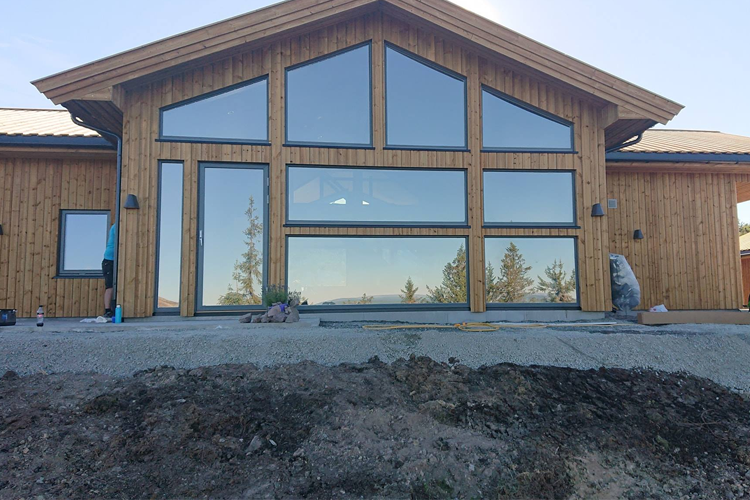 Moderne hytte med store vinduer og trepanel, omgitt av natur og fjellutsikt.