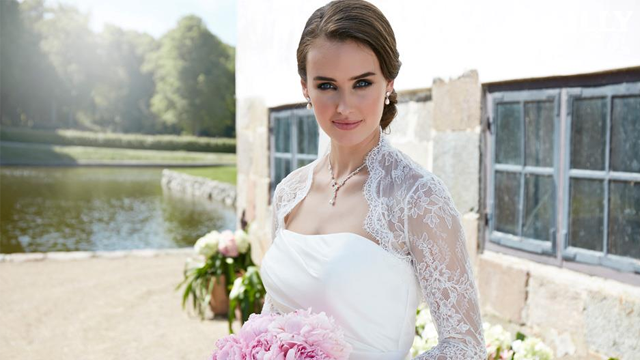 Brud med blomsterbukett står ved vannet, iført en elegant brudekjole med blonde og perler.