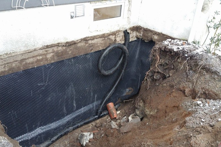Gravd ut område rundt en bygning med svart dreneringsnett og rør synlig i jorden.
