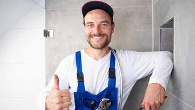 En smilende rørlegger i blå arbeidsklær gir tommel opp med verktøy i hånden.