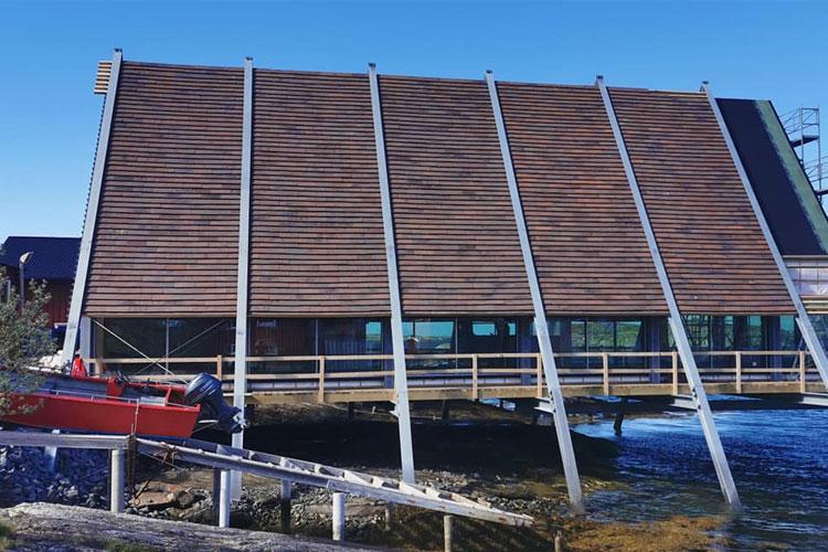 Moderne bygning med skrå tak og store vinduer, plassert ved vannkanten med en båt på en rampe.