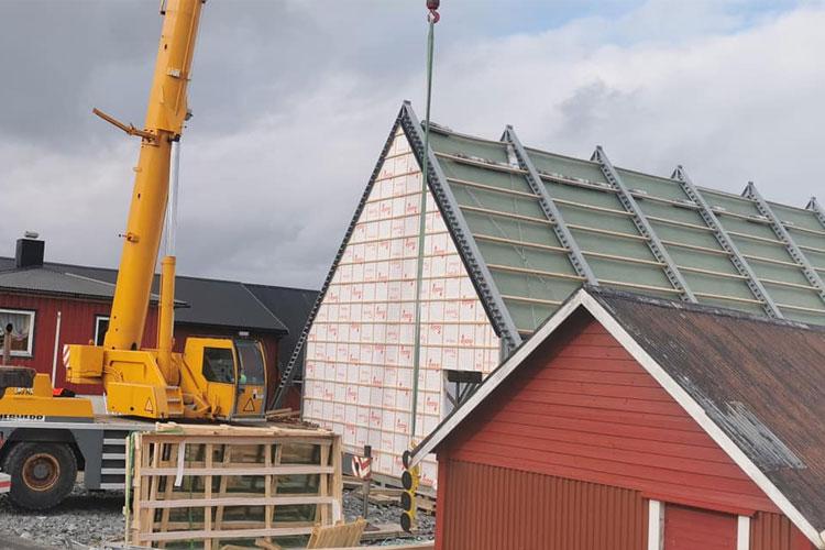 Bygging av et hus med kran som løfter takplater, med rød bygning i forgrunnen.