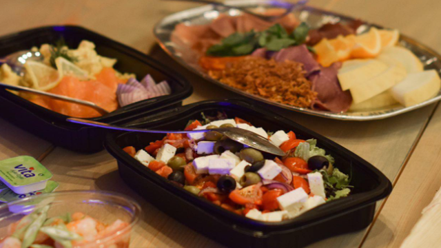 Fargerik salat med fetaost, oliven og cherrytomater, servert med skinke og frukt på et bord.