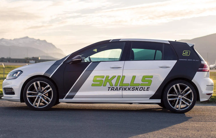 En bil med "Skills Trafikkskole" på siden, parkert ved en vei med fjell i bakgrunnen.