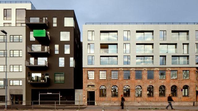 Moderne leilighetsbygg i svart og grått ved siden av en gammel mursteinsbygning med store vinduer.
