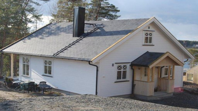 En moderne enebolig med grå takstein, hvit fasade og veranda, omringet av trær og grus.