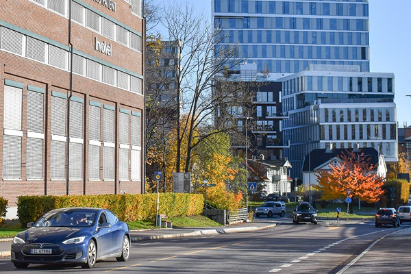 En blå bil kjører forbi et hotell og moderne bygninger med høstfarger i trærne langs veien.