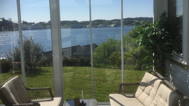 Utsikt fra en solfylt veranda med to stoler, et bord og en vakker sjøutsikt. Grønne trær og båter i bakgrunnen.