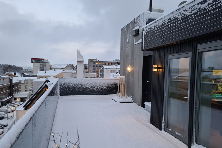 En snødekt takterrasse med utsikt over bygninger og en grå himmel. Snøen dekker gulvet og rekkverket.