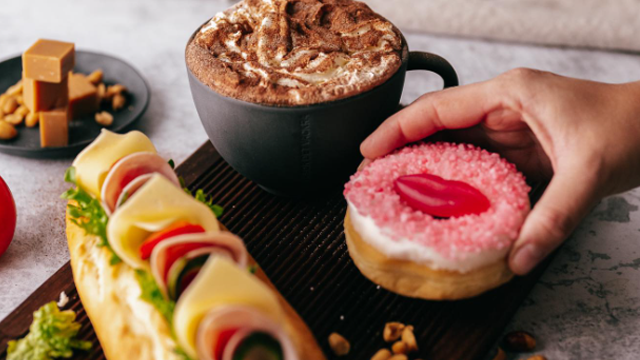 En hånd tar en rosa donut med gelé, ved siden av en sandwich med ost og grønnsaker, og en kopp kakao med krem.