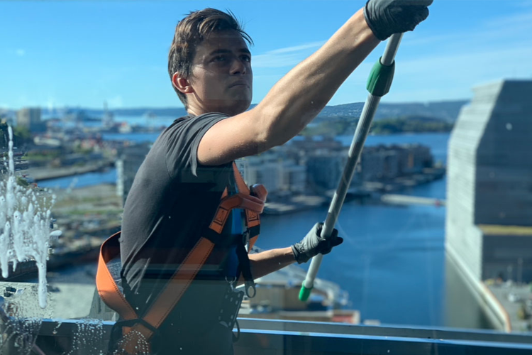 En vindusvasker rengjør vinduet på en høy bygning med utsikt over byen og vannet.