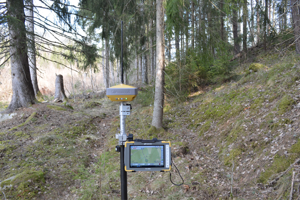 GPS-utstyr med skjerm i skogen, omgitt av trær og mose, brukt til kartlegging og måling.
