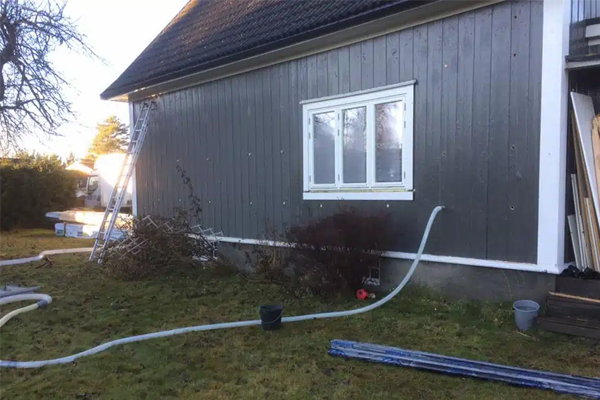 Husvegg med grå panel, vindu, rør og verktøy i hagen, samt en stige og busker i bakgrunnen.
