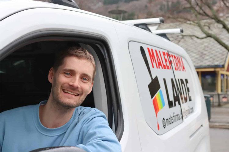 Mann smilende fra vinduet i en hvit varebil med malerfirmaets logo og kontaktinformasjon.