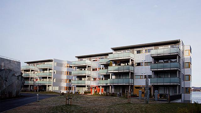Moderne leilighetsbygg med balkonger og grøntområde foran, beliggende ved en vei.