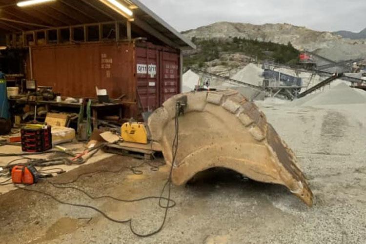 En stor gravemaskin skuff ligger på bakken i en steinbrudd, med verktøy og utstyr rundt.