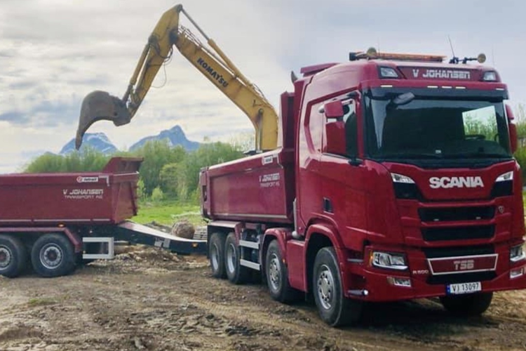 En rød Scania lastebil med gravemaskin som laster jord i en tilhenger på en byggeplass.