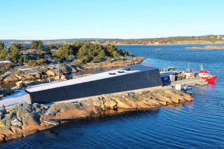 Moderne bygning ved vannkanten med en rød båt og steinete kystlinje i bakgrunnen.