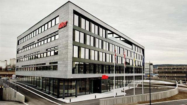 Moderne kontorbygg med store vinduer og rød logo på fasaden, beliggende i et urbant område.