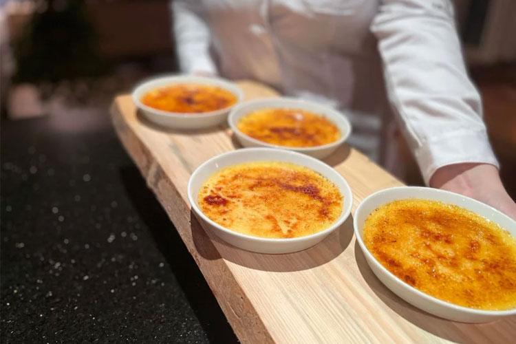 Fire porsjoner crème brûlée servert på en treplate, med en person som holder brettet.