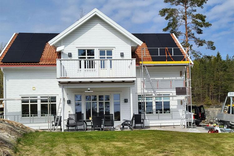 Moderne, hvitt hus med solcellepaneler, balkong og stillas i hagen, omgitt av trær og blå himmel.