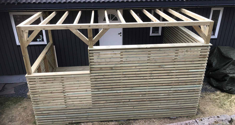 Trekonstruksjon med takstoler og vegger av tre, delvis ferdig bygget ute ved en bygning.