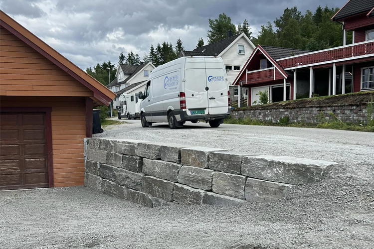 En grå steinmur ved en grusvei med en varebil som kjører forbi, omgitt av hus og trær.