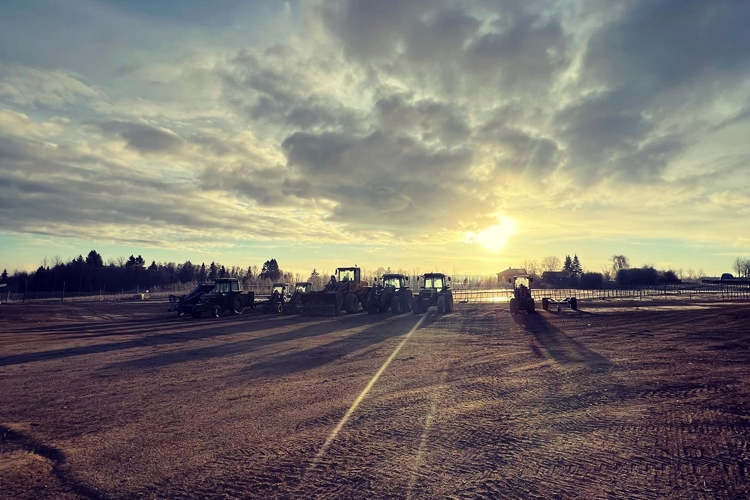 Traktorer står parkert på en åpen mark med solnedgang og skyer i bakgrunnen.