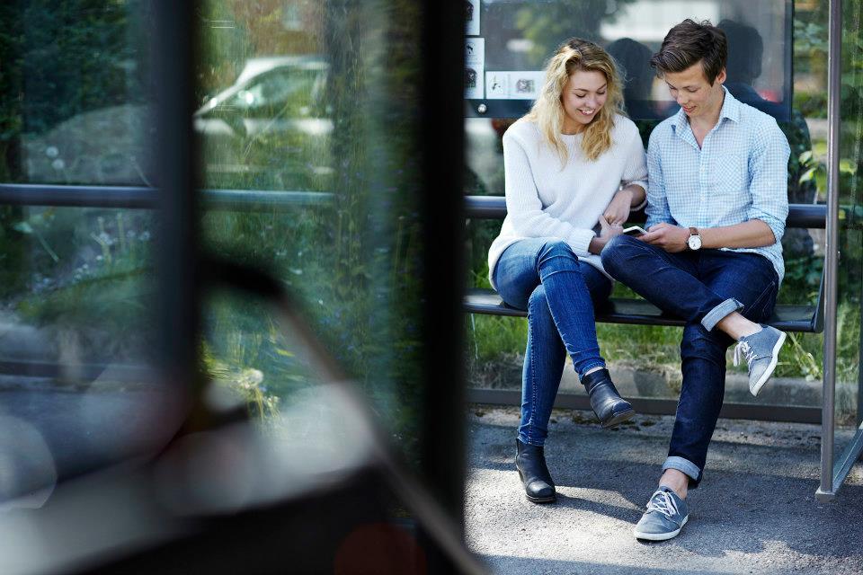 Unge par sitter på en benk på busstopp, smilende og ser på telefonen sammen. Solfylt dag med grønn bakgrunn.