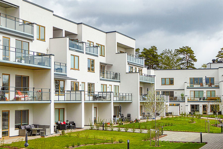 Moderne leilighetskompleks med balkonger og grøntområde, omgitt av trær og en skyet himmel.
