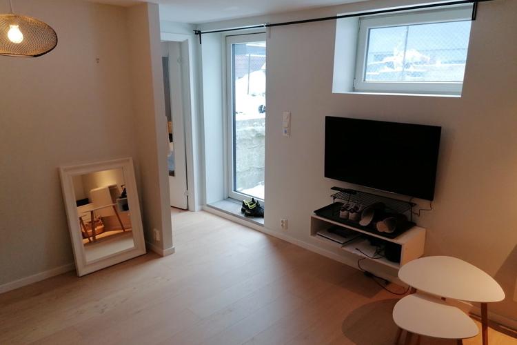 Lys stue med TV, speil og vindu som gir utsikt til utsiden. Møbler i moderne stil.