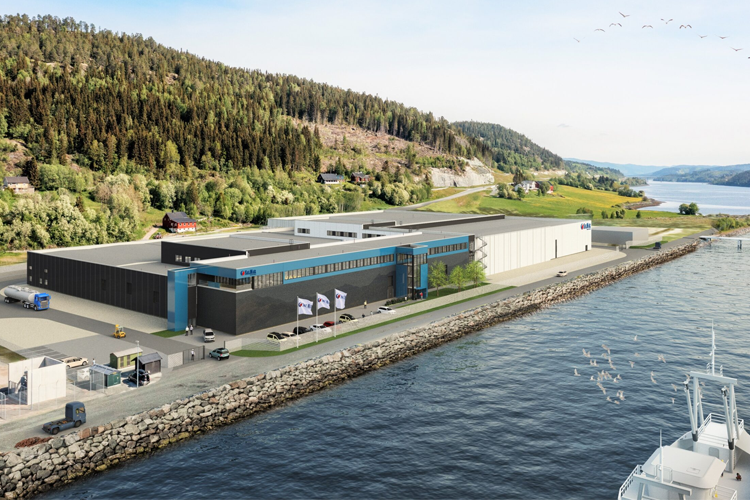 Moderne fabrikkbygning ved vannkanten, omgitt av skog og fjell, med biler og båter i nærheten.