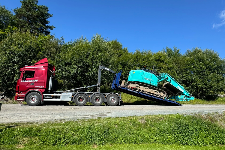 En lastebil med en grønn gravemaskin på en tilhenger, parkert langs en grusvei omkranset av trær.