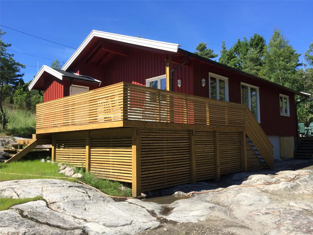 Rødt hus med terrasse i tre, omgitt av grønn natur og steinlandskap. Solfylt dag med blå himmel.